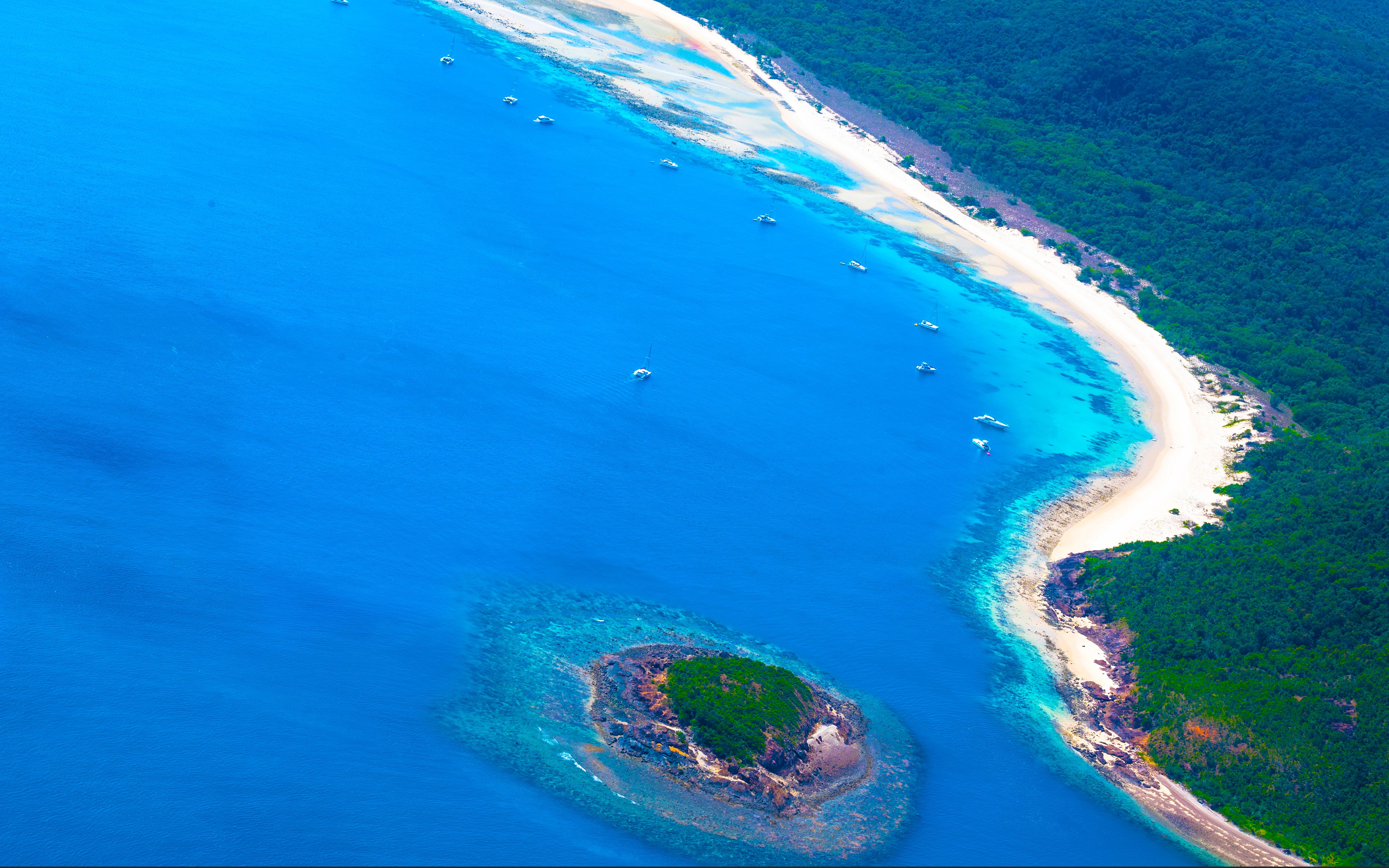 Aerial view of a turquoise bay with boats at anchor along a white sand beach backed by forest.