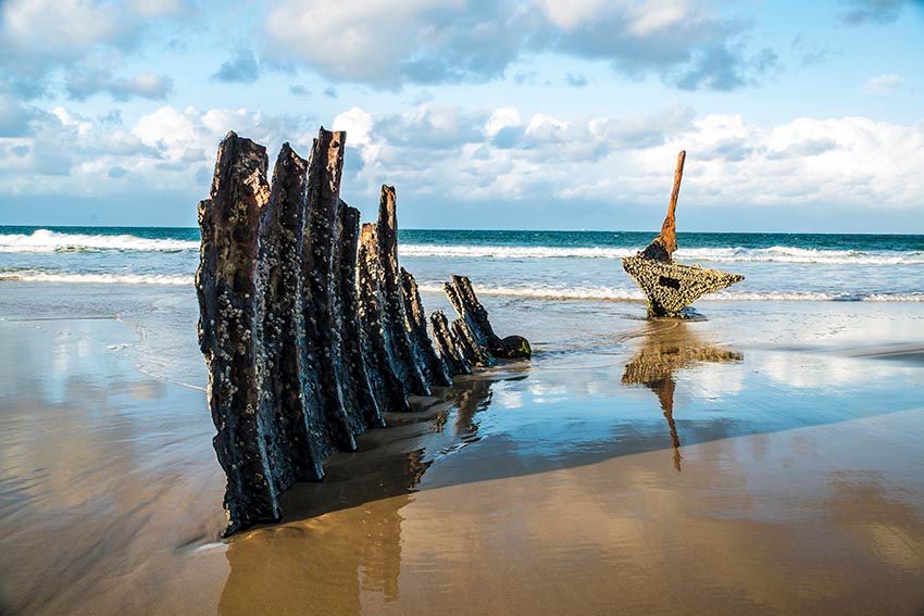 Weathered wooden shipwreck timbers standing on the sand at Dicky Beach, with waves breaking behind.