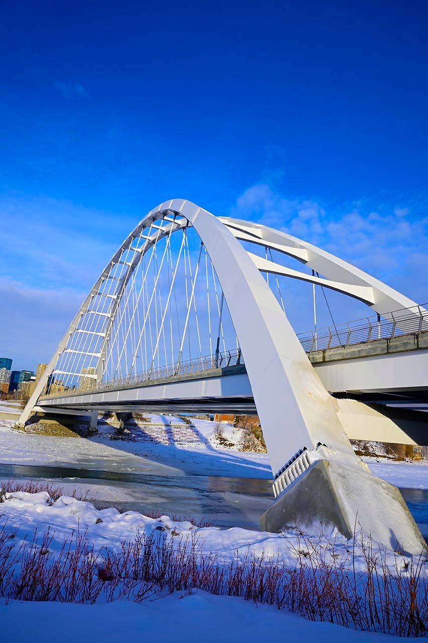 A white arched pedestrian bridge over a frozen, snow-lined river in Edmonton, Canada.