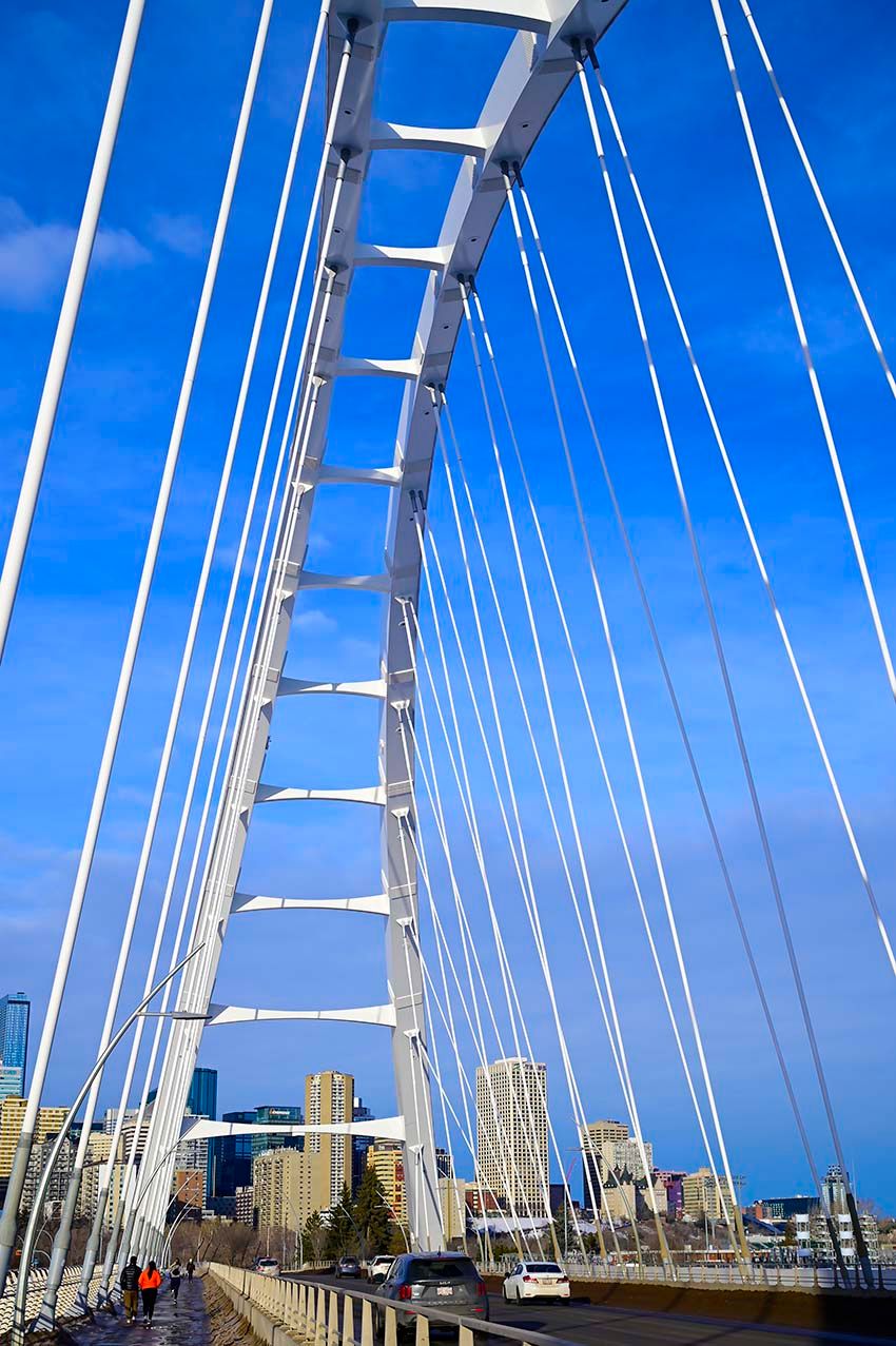 A cable-stayed pedestrian bridge tower against a blue sky above the Edmonton, Canada skyline.