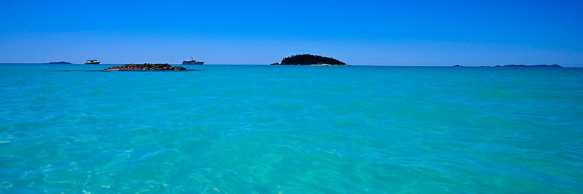 A wide panorama of turquoise-to-deep-blue water with a small forested island and moored boats on the horizon.