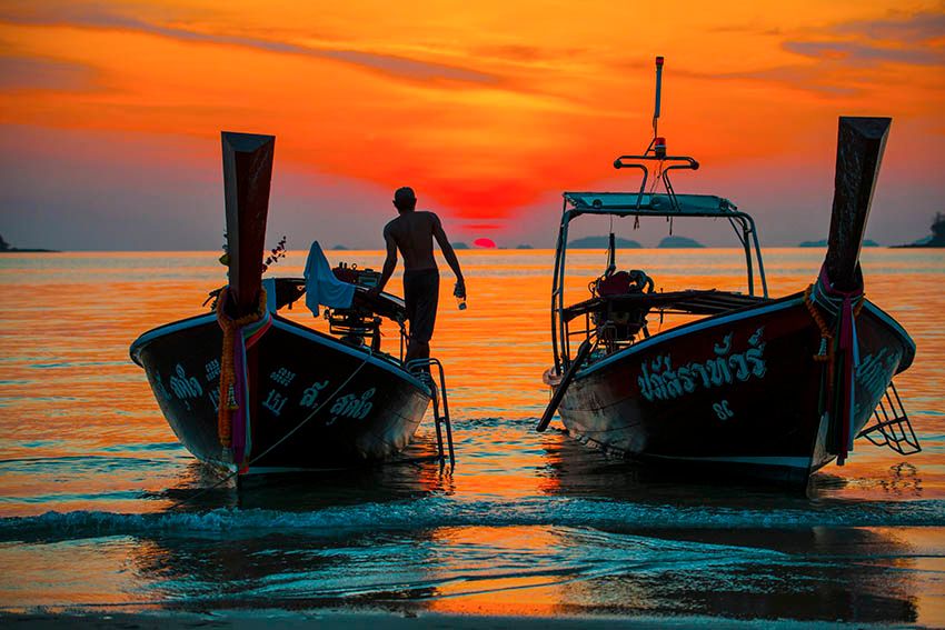 Silhouette of a man standing between two traditional longtail boats on a beach at a fiery sunset.