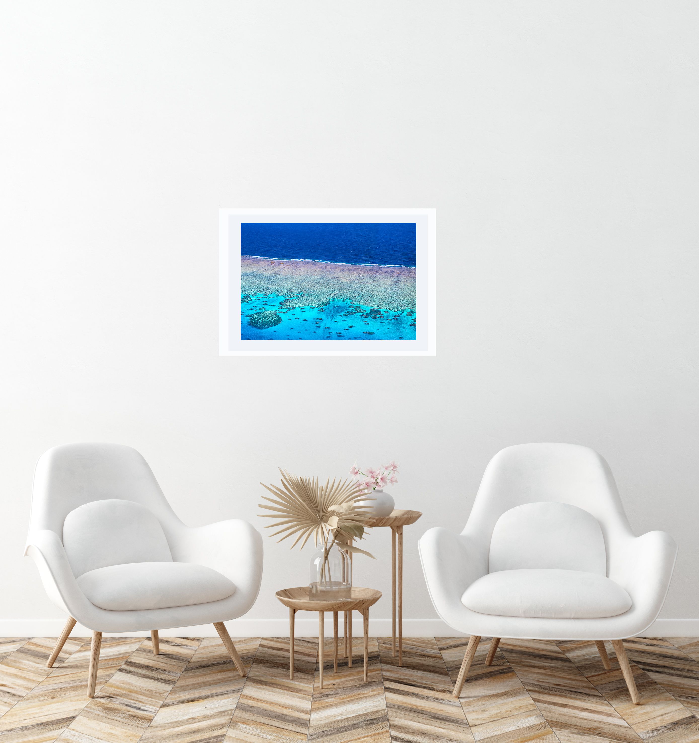 An aerial reef photo in a white frame with mat, showing turquoise water and coral over a beach edge, hung above a side table.