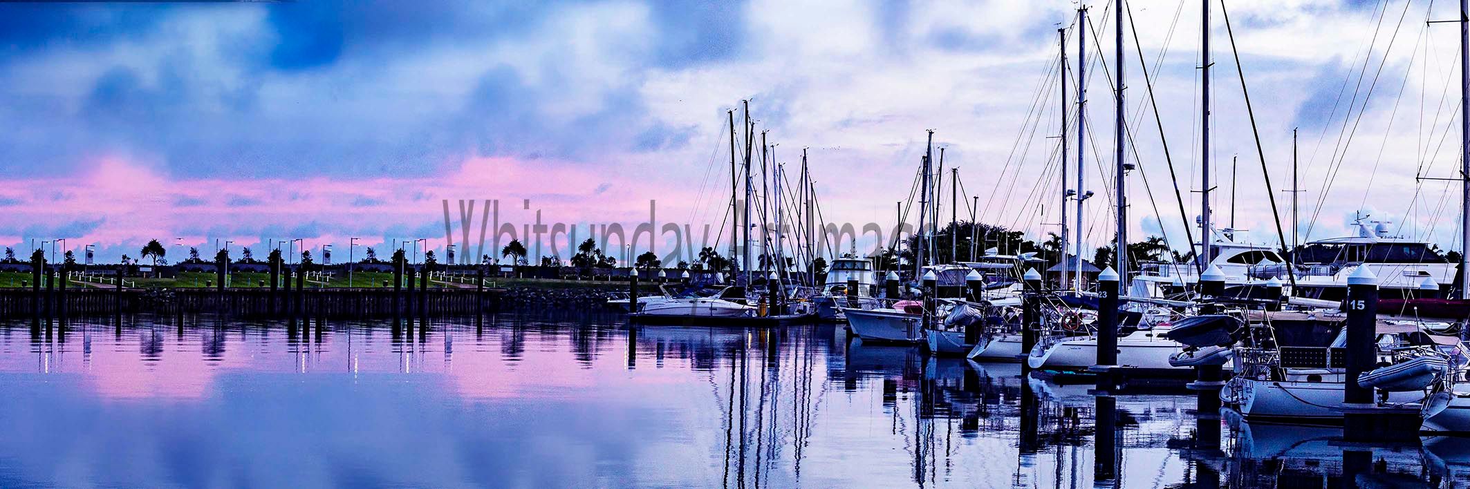 A wide panoramic view of yachts moored at the Port of Airlie marina under a pink and blue sunrise sky.