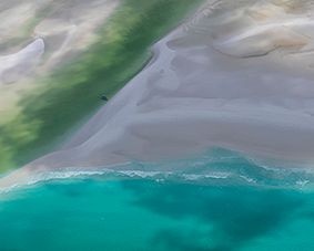 An aerial view of pale sand and shallow water meeting deeper turquoise sea in soft, painterly bands.