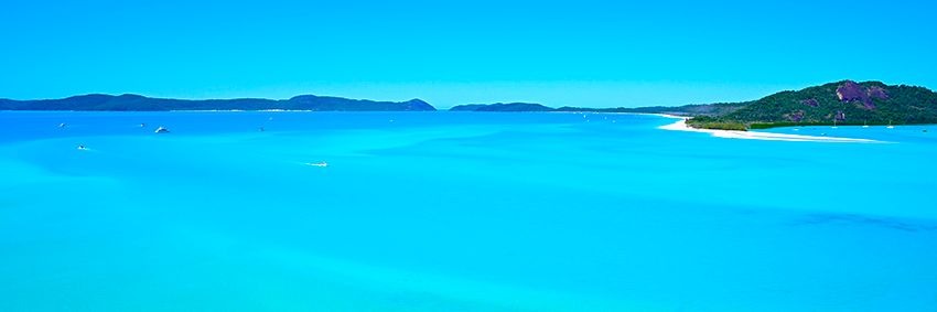 A wide panorama of vivid turquoise water and white sand meeting a forested headland under a clear sky.